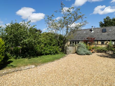 A garden with a gravel area and trees next to a stone house with a slate roof at Henmarsh Cottage in Moreton-In-Marsh