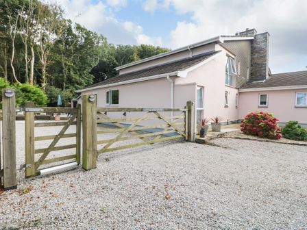 A gravel driveway with a wooden gate in front of a house with pink exterior walls and a stone chimney at Little Westwards in Goldsithney