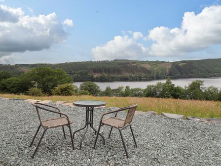 Two chairs and a small round table on gravel with a lake and forested hills in the background at Llety Dewi in Lake Vyrnwy