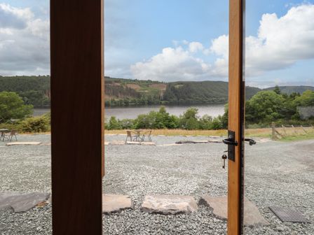 A view through wooden framed glass doors onto a gravel area with outdoor tables and chairs by a lake with hills and trees at Old Cart House in Llanwddyn