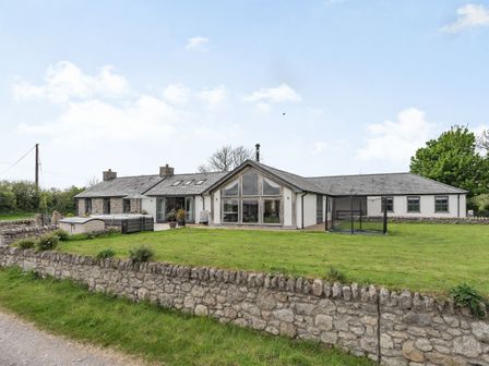 A house with large windows a stone wall a trampoline and a hot tub in the yard at Hen Hafod in Talwrn near Llangefni