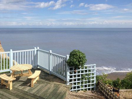 A wooden deck with a round table and two benches overlooking a calm sea with a white railing and some plants at Anchorage in Watchet