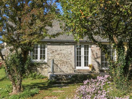 A stone cottage with white framed windows and a door with two steps and trees and flowers outside at The Daffodil Shed in Bere Alston