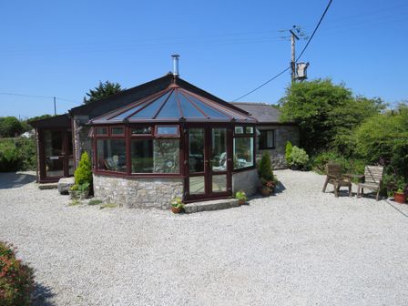 A stone house with a glass conservatory and chairs outside on a gravel area at Sheilas Cottage in Penryn