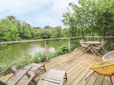 A wooden deck with lounge chairs a round table with chairs and a yellow chair overlooking a river surrounded by trees at 2 Water's Edge Lanreath