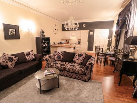 A living room with patterned sofas a glass coffee table a chandelier a dining table and kitchen area at Sneaton Hall Apartment 4 in Sneaton