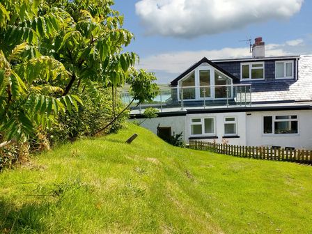 A white house with glass balcony on a hill with grass and trees at Waters Edge in Dale