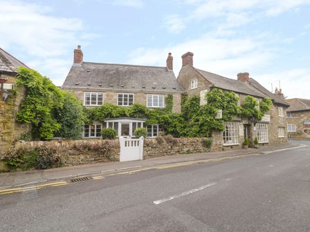 Stone buildings covered with greenery along a street with a white gate at Market House in Abbotsbury