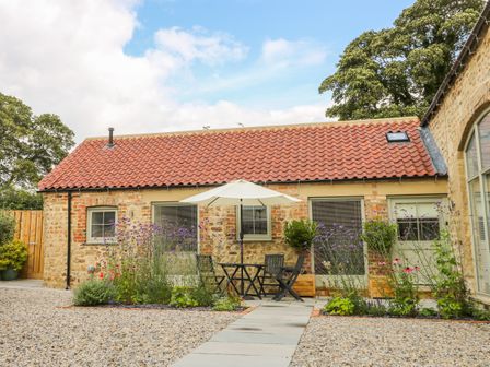 A stone cottage with red tiled roof and a patio with table umbrella and chairs surrounded by flowers at Wallerthwaite Barn Cottage in Markington