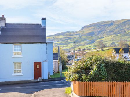 A street with a blue house with a brown door and windows next to a hedge with a wooden fence and hills in the background at Cranny Corner in Carnlough