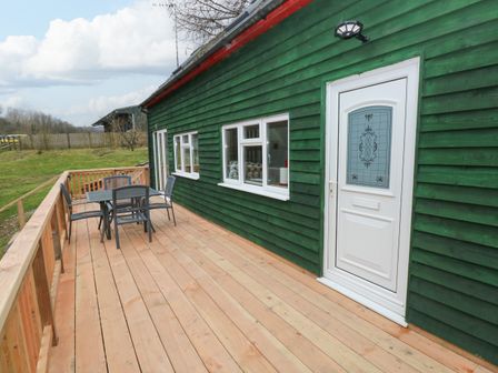 An outdoor wooden deck with a table and four chairs next to a green wooden building with windows and a white door at The Hen Pen in Star near Cenarth