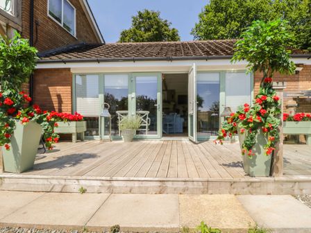 A wooden deck patio with green planters and red flowers in front of glass doors at High Rigg Woodland Cottage in Brampton Cumbria