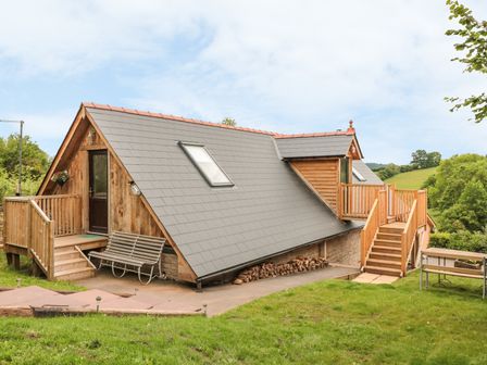 A wooden house with a grey sloped roof and stairs with railings surrounded by grass and trees at The Lookout in Llangrove