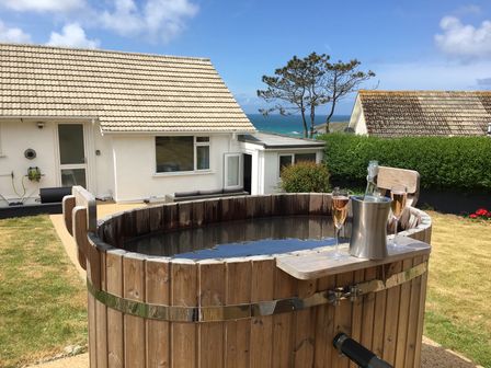 A wooden hot tub with two glasses of rose wine and a wine bottle in a cooler on a lawn with houses and ocean in the background at Chi An Mor in Porth
