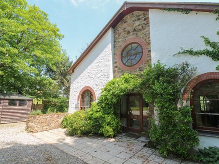 An exterior view of a stone and white plaster house with arched windows and a wooden door surrounded by greenery and a gravel pathway at Beggars Roost in Penhallow