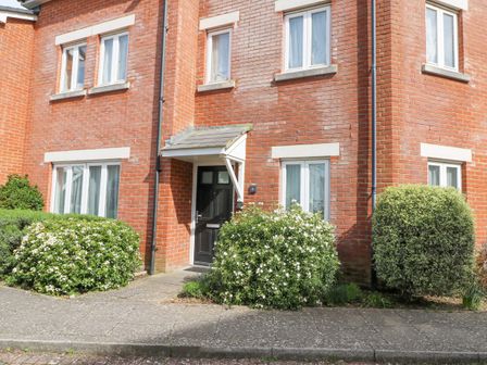 The front exterior of a brick house with a black door and white framed windows surrounded by green bushes at Admirals Quarter Apartment 4 in Weymouth