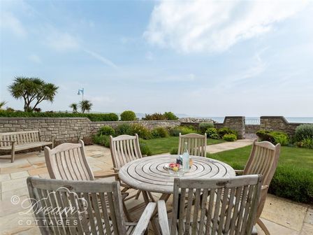 An outdoor patio with a round wooden table and six chairs next to a garden and stone wall overlooking the sea at Beach View Apartment 2 in Greenhill
