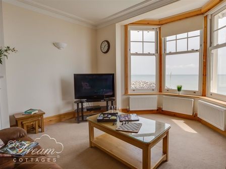 A living room with a TV on a stand a coffee table with board games a clock on the wall and large windows showing a sea view at Beach View Apartment 3 in Greenhill