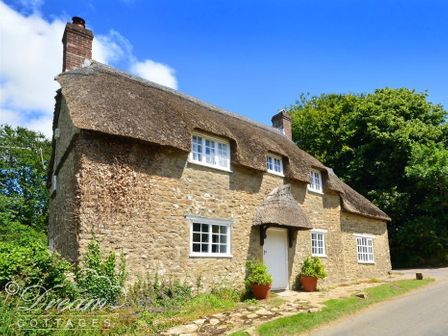 A stone cottage with a thatched roof and white windows beside a road at Little Berwick Cottage in Burton Bradstock