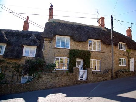A row of stone cottages with thatched roofs and white framed windows along a sloped street at Bramble Cottage in Burton Bradstock