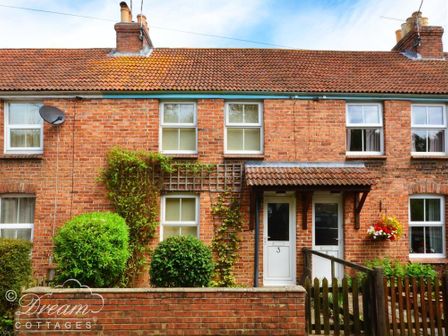 A brick cottage with two white doors numbered 2 and 3 surrounded by green bushes and flowers at Brickyard Cottage in Moreton