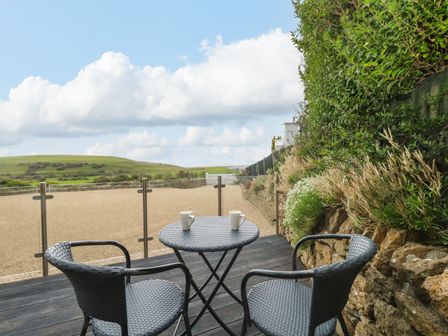 A patio with a round table and two chairs with two mugs on the table overlooking a gravel area and green hills at Burton Bay in Burton Bradstock