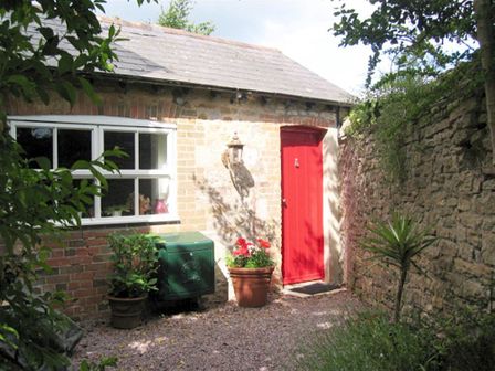 An outside view of a small brick building with a red door and a window with plants and a green utility box at Fountain Cottage in Weymouth