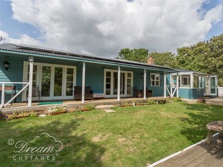 A garden with grass and flowerbeds in front of a blue bungalow with a veranda and outdoor chairs at Harbour View Bungalow in Weymouth