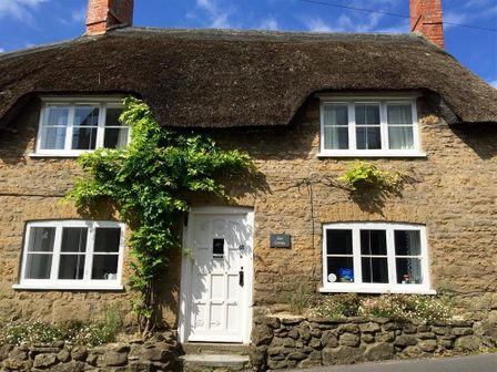Exterior of a stone cottage with a thatched roof white door and windows with plants around the entrance at Lilac Cottage in Burton Bradstock