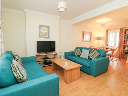 A living room with teal sofas a wooden coffee table a television on a corner stand and a dining area with wooden chairs at Louisa Cottage Brewers Quay Harbour
