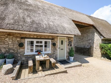 A stone cottage with a thatched roof and a wooden table and benches outside at Magnolia Cottage Osmington