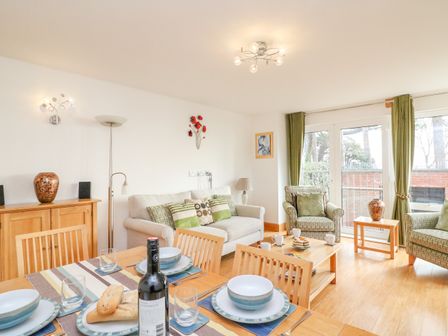 A dining table set with plates glasses and bread next to a living room with sofa armchairs coffee table and glass doors with green curtains at Newton's Cove View in Weymouth