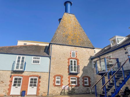 A stone building with a conical roof and blue chimney pipes next to brick and blue wooden sections at The Oast House in Brewers Quay Harbour