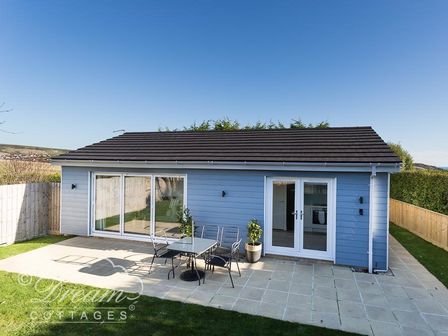 A small blue house with patio furniture and potted plants on a tiled patio at Outlook Lodge in Bowleaze