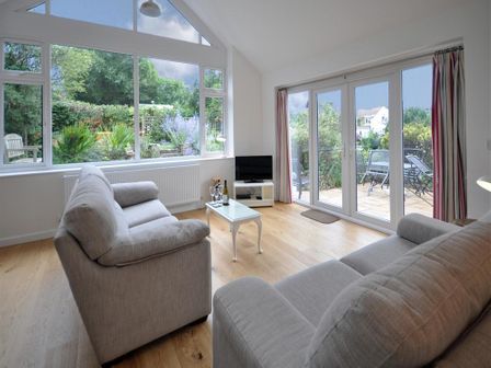 A living room with two sofas a small white table with wine glasses and bottle a tv and glass doors to a garden at Pebbles in Burton Bradstock