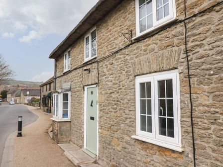 A stone cottage with white framed windows and a light green door along a street in a village