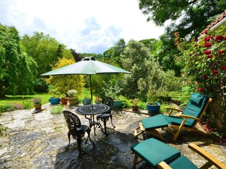 A patio with a round table and chairs under a green umbrella surrounded by potted plants and garden furniture at Sixpenny Cottage in Upwey