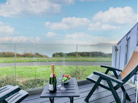 A balcony with two chairs a table with a bottle of champagne and glasses overlooking greenery and water at Waters Edge in Wyke Regis