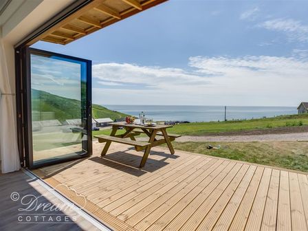 A wooden deck with a picnic table set with glassware and fruit overlooking a grassy area and the sea at The Wave in Eype
