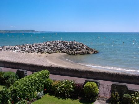 A rocky jetty and small sandy beach by the sea with a garden and gated walkway in the foreground at Weymouth Bay Apartment B in Greenhill
