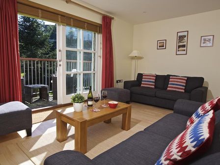 A living room with two gray sofas with patterned pillows a wooden coffee table with wine glasses and a plant and sliding glass doors leading to a balcony at 1 Combehaven in Salcombe