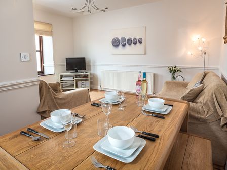 A dining table set with plates glasses and cutlery in a living room with covered sofas a television and a floor lamp at 2 Armada House in Hope Cove