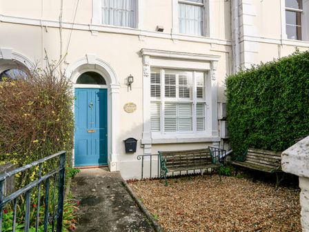 The exterior entrance of a beige house with a blue door two benches on gravel and hedges on either side at 2 North Crest House in Salcombe