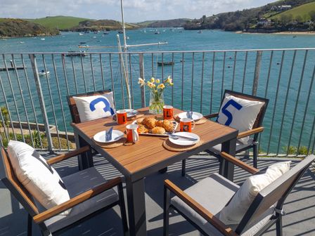 A balcony dining table set for four with pastries and coffee mugs overlooking a body of water with boats at 35 The Salcombe in Salcombe