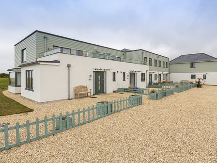 A modern two-story building with small fenced garden areas and a bench outside at 3 Ocean Reach in Bolberry