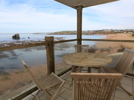 A balcony with a wooden table and four chairs overlooking a rocky beach and ocean at 4 Thurlestone Rock in Thurlestone