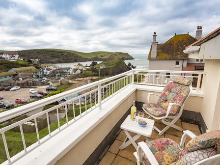 A balcony with two cushioned chairs a small table with a bottle glass and bowl overlooking a coastal village and green hills at 5 Chichester Court in Hope Cove