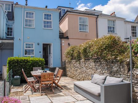 A patio with a wooden table and chairs and a grey outdoor sofa in front of pastel-colored houses at Bumblebee Cottage in Salcombe