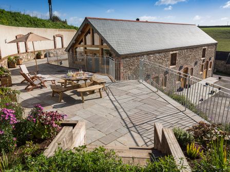 An outdoor patio area with a wooden picnic table set with plates and drinks an umbrella and chairs near a stone building with a tiled roof at Butterwell Barn in Dartmouth