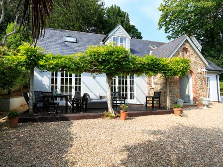 An outdoor patio with black chairs and tables under a pergola with greenery in front of a house with stone and brick facade at The Chota House in Salcombe